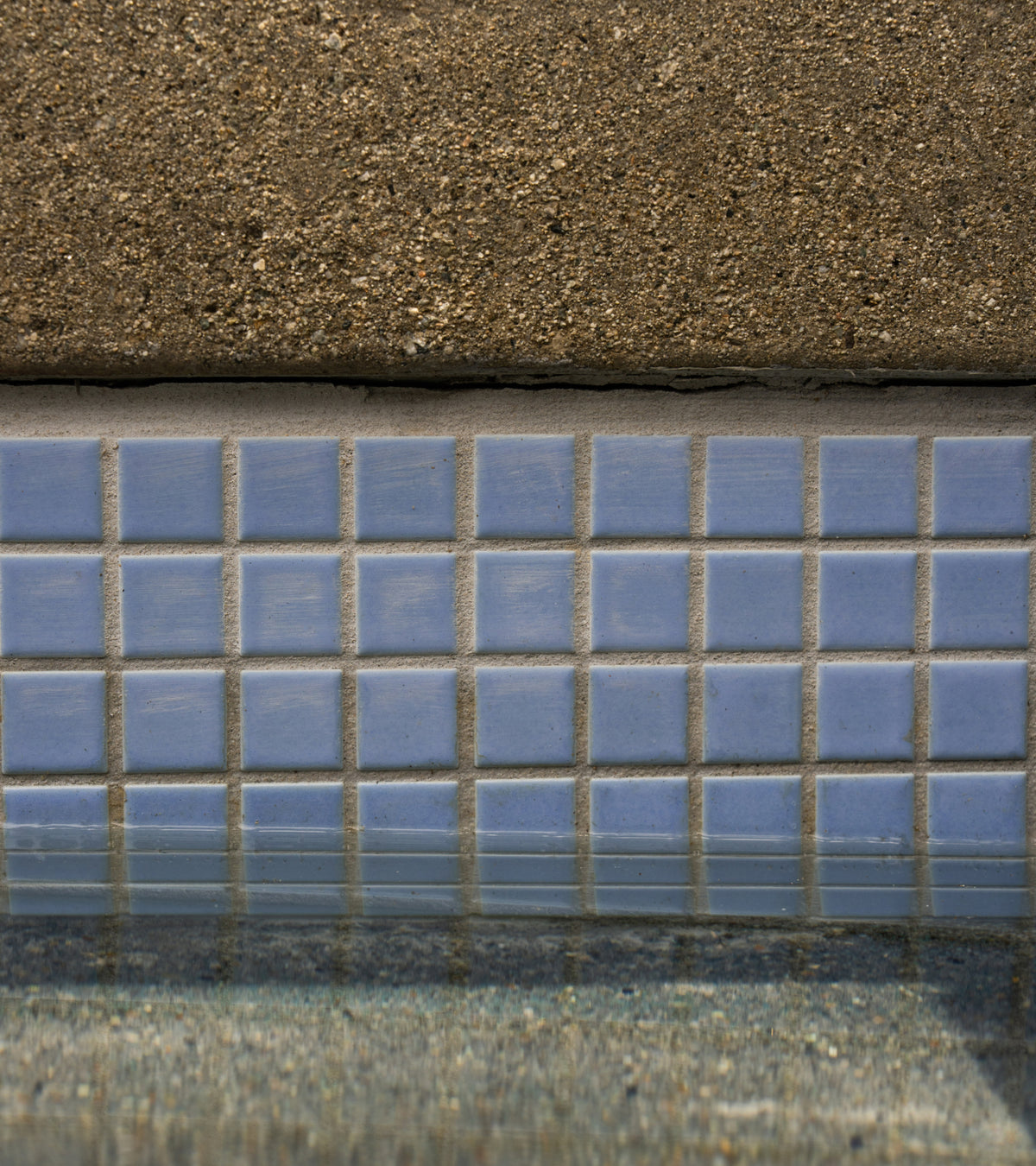 A row of Cornflower 1x1 light blue square tiles bordering the edge of a swimming pool below a concrete deck.