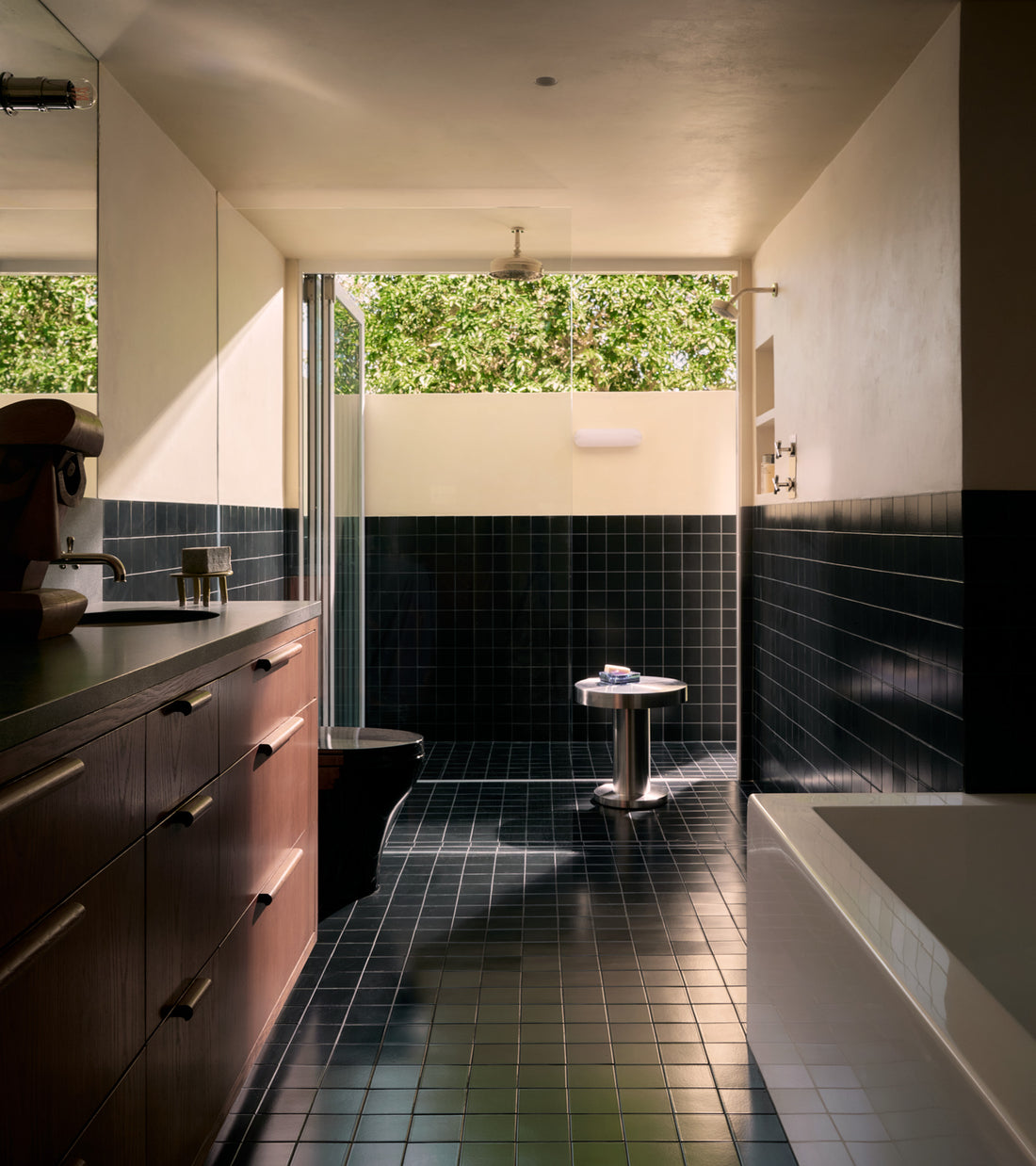 Sunlit modern bathroom with Zia Tile's Cosmic Black 4x4 ceramic tiles on floor and walls, wooden cabinets left, walk-in shower at the back, white bathtub right, and a large window showing greenery outside.