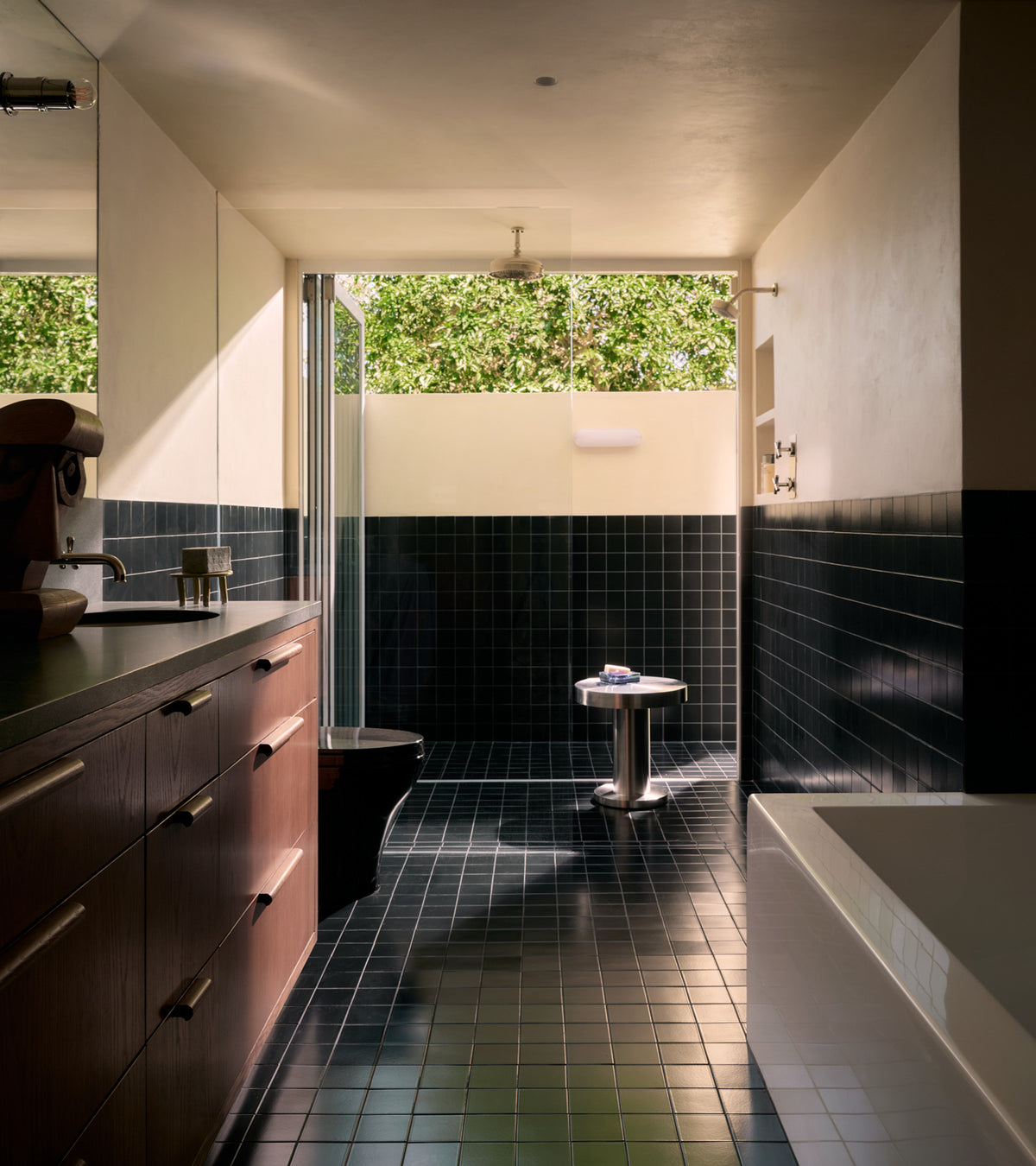 Sunlit modern bathroom with Zia Tile's Cosmic Black 4x4 ceramic tiles on floor and walls, wooden cabinets left, walk-in shower at the back, white bathtub right, and a large window showing greenery outside.