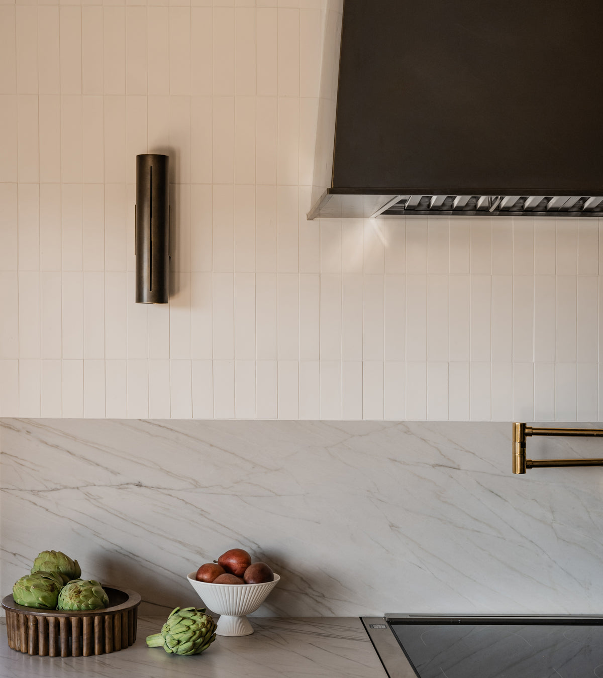 A modern kitchen with a marble countertop, artichoke and apples on the counter, a ribbed bowl, a Cotton 2x8 backsplash by vendor-unknown, brass pot filler, and stainless steel range hood above.