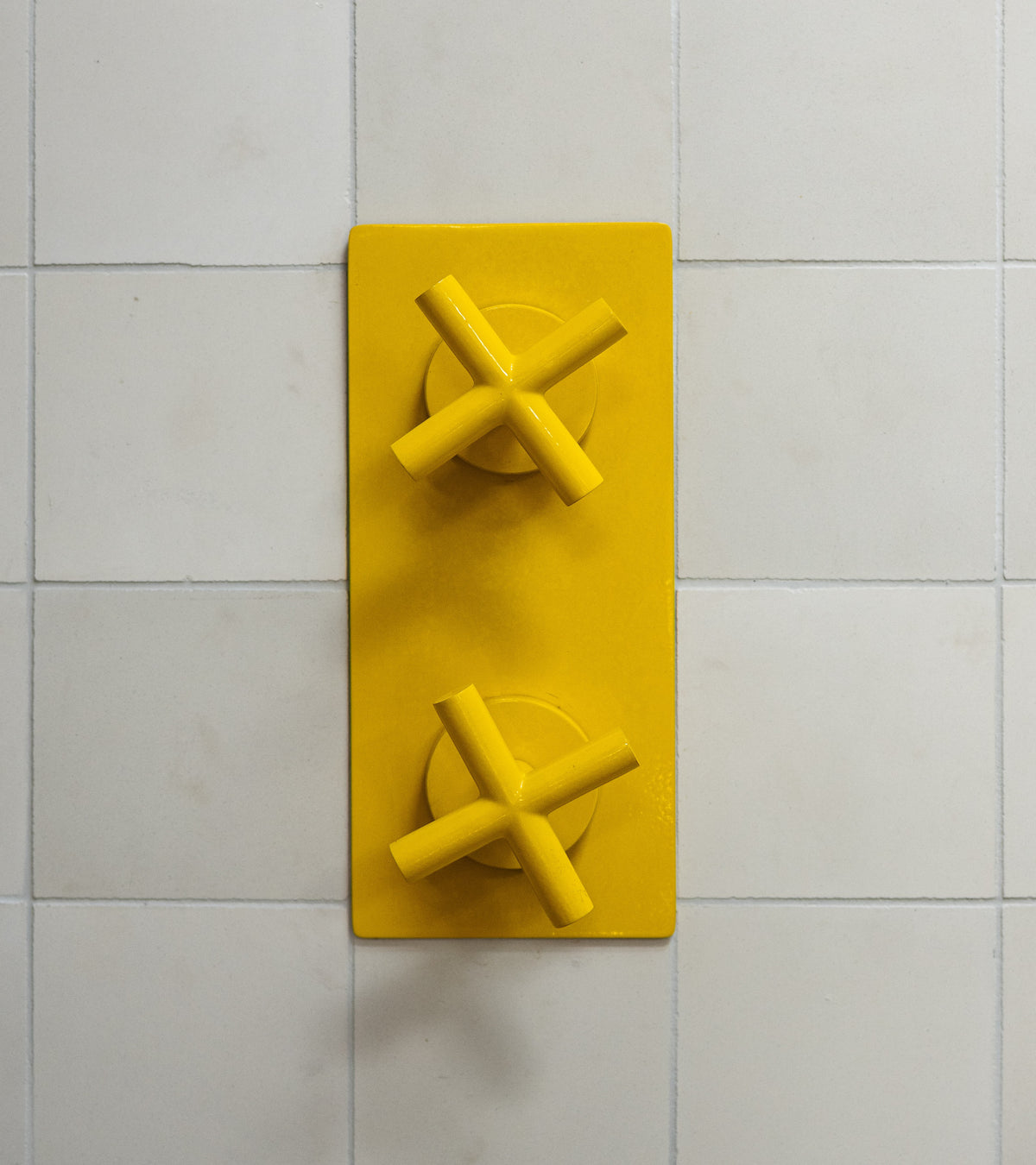 Two bright yellow, X-shaped knobs from vendor-unknown's Cotton 4x4 are mounted vertically on a rectangular yellow plate against white tiled backsplash, viewed from above to emphasize the geometric composition.