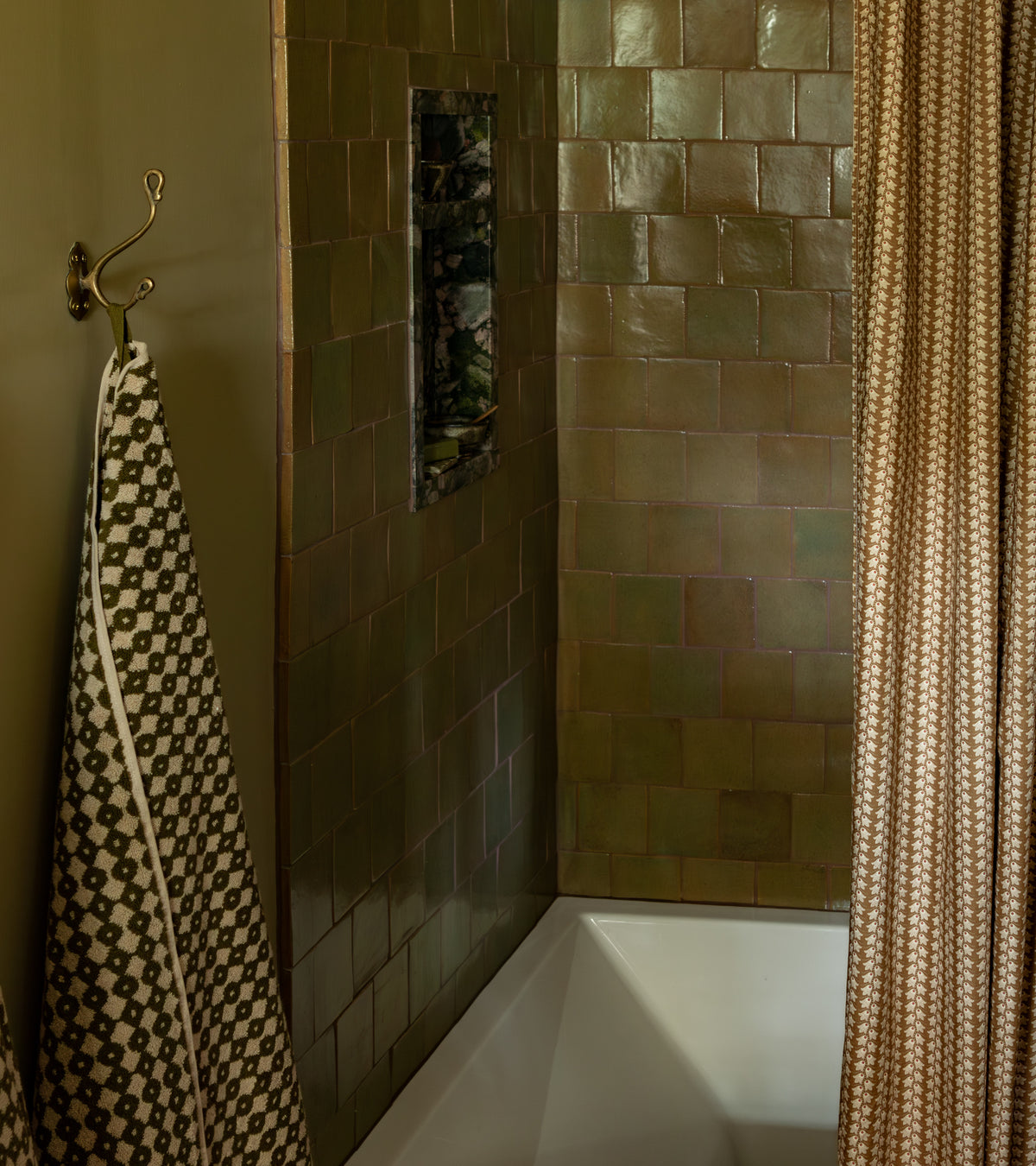 A bathtub with Creosote 4x4 Square handmade olive green tiles by Zia Tile on the shower walls, a small built-in shelf, a patterned towel on a hook, and a partially closed beige patterned shower curtain.