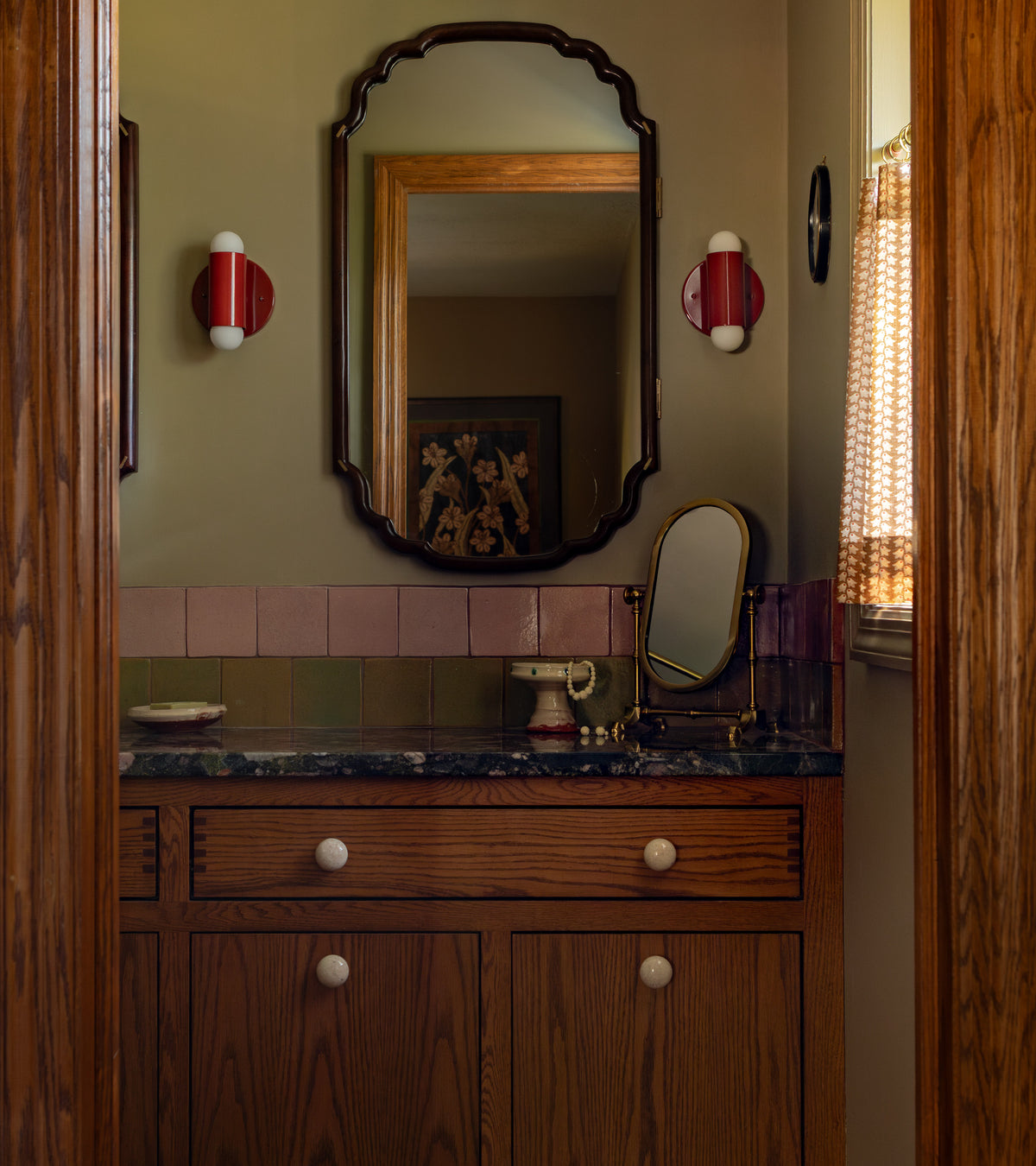 A cozy bathroom with wooden cabinets, marble countertop, scalloped wall mirror, red and white sconces, a vintage mirror, and patterned curtains features handmade Creosote 4x4 Square tiles by Zia Tile inspired by ceramic arts.