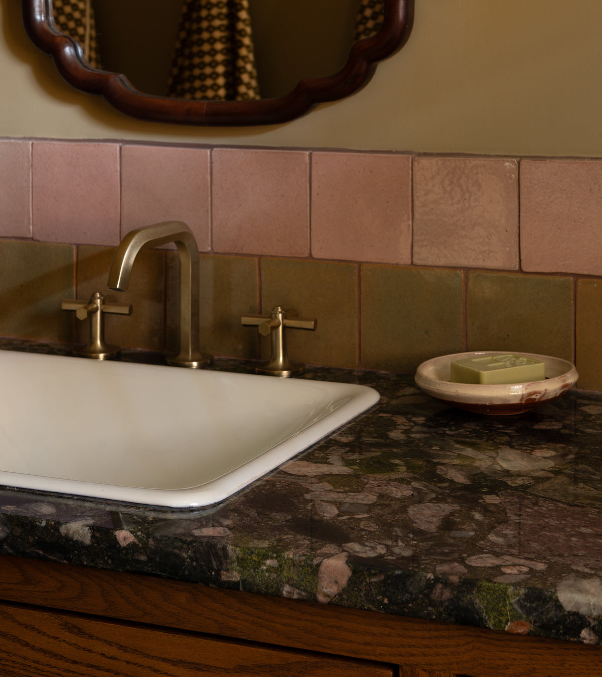 A bathroom sink with a marble countertop, brass two-handle faucet, and a handmade Cotto soap dish with green soap is set against a pink and olive Posada 4x4 Square Zia Tile backsplash; a wood-framed mirror completes the look.