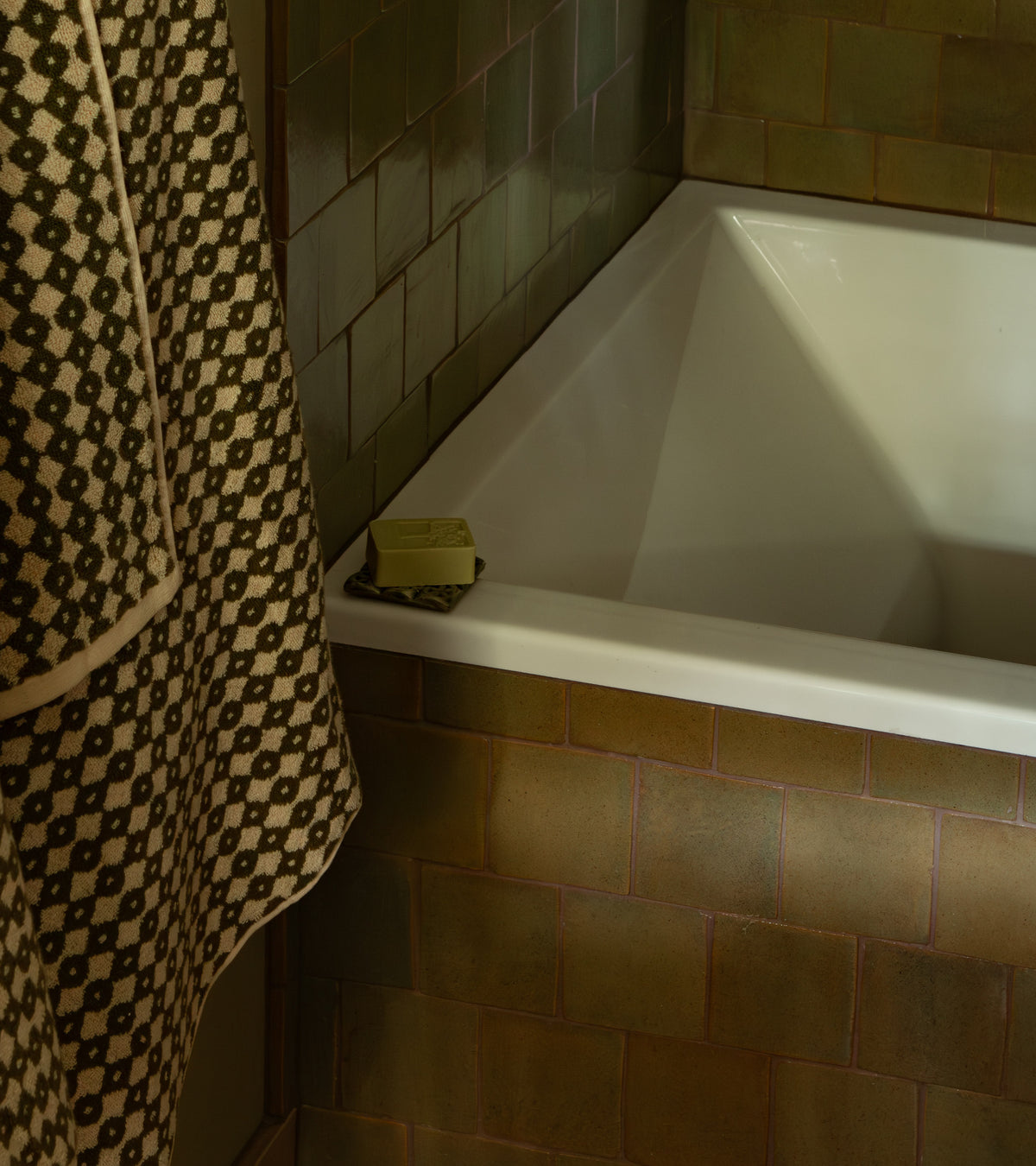 A white bathtub corner features Zia Tile's Creosote 4x4 Square tiles in olive green, a bar of soap on the tub edge, and a geometric towel nearby, creating an artisanal atmosphere.