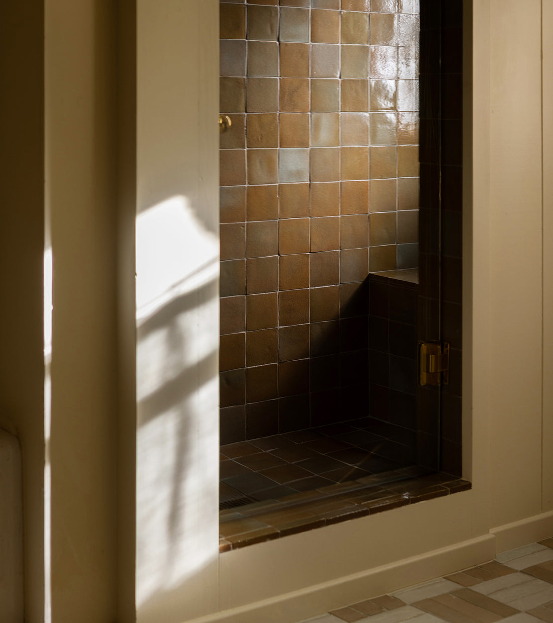 A shower featuring Zia Tile's Creosote 4x4 Square brown tiles and a built-in bench, with sunlight streaming through a nearby window onto the wall and tiled floor, showcasing timeless ceramic craftsmanship.