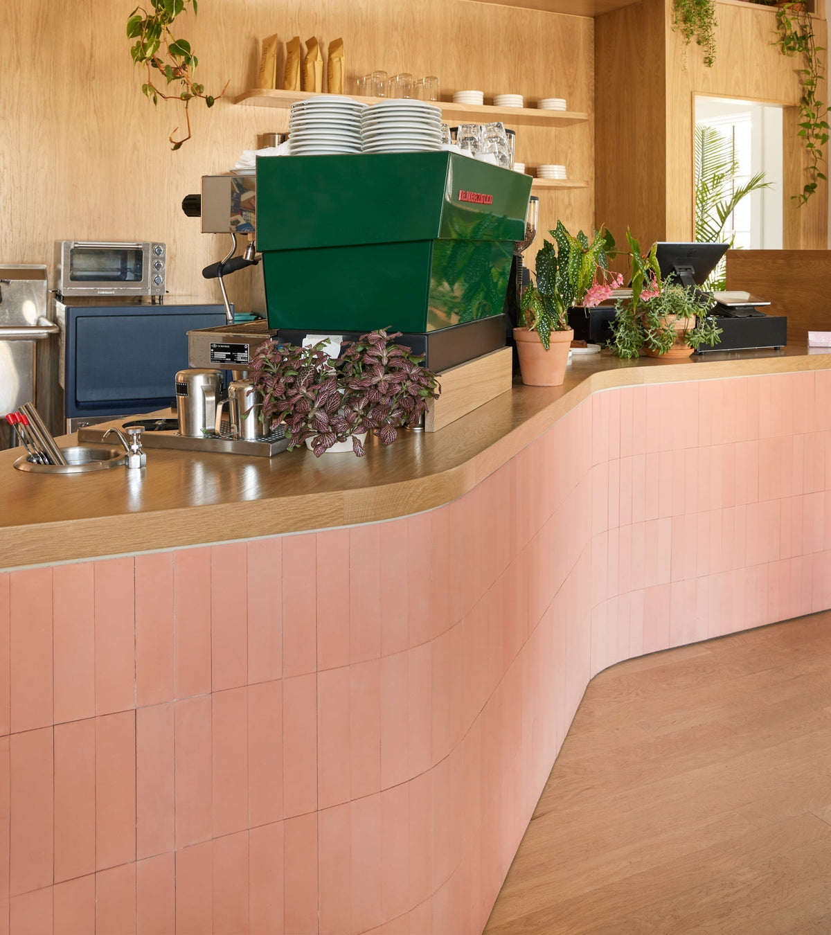 A modern café counter features a curved handmade pink Delta Moon 2x8 tile front by vendor-unknown, a green espresso machine, potted plants, and wooden shelves with cups and plates in a warm wood-paneled setting.