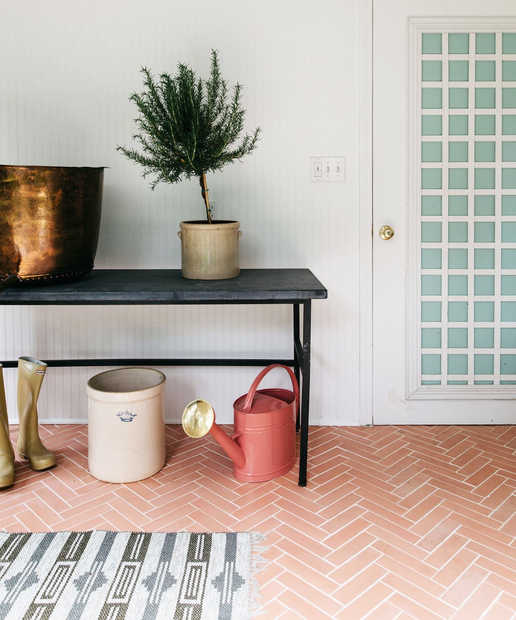 A black table with a potted plant and copper tub sits against a white wall. Yellow rain boots, a large crock, and pink watering can are on handmade tile flooring featuring Delta Moon 2x8 from vendor-unknown. A glass grid door is on the right.