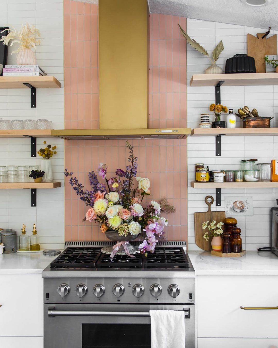 A modern kitchen features a stainless steel stove, Delta Moon 2x8 handmade tile backsplash by vendor-unknown, and a gold range hood. Fresh flowers decorate the stove, with open shelves showing dishes, glass jars, and decor.