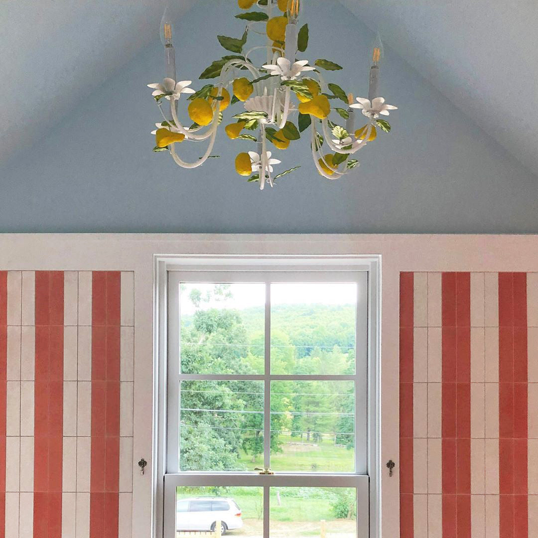 A white-framed window with a green landscape sits between pink-striped walls. Below, Delta Moon 2x8 tiles from vendor-unknown add character, while a lemon and leaf chandelier brings a whimsical touch above.