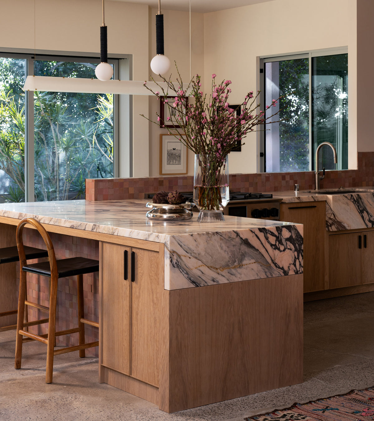 A modern kitchen with light wood cabinets and large windows features an elegant backsplash of Desert Bloom 2x2 tiles by Zia Tile, complementing the marble island, black chair, pink flowers in a vase, and pendant lights.