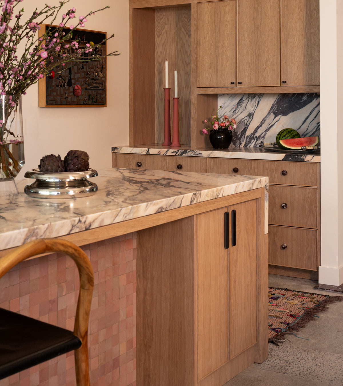 A modern kitchen features light wood cabinets, marble countertops, a fruit bowl, candles, flowers in a vase, and a rug. The island's side is accented with Desert Bloom 2x2 tiles from Zia Tile for Moroccan flair. A black chair is partially visible.