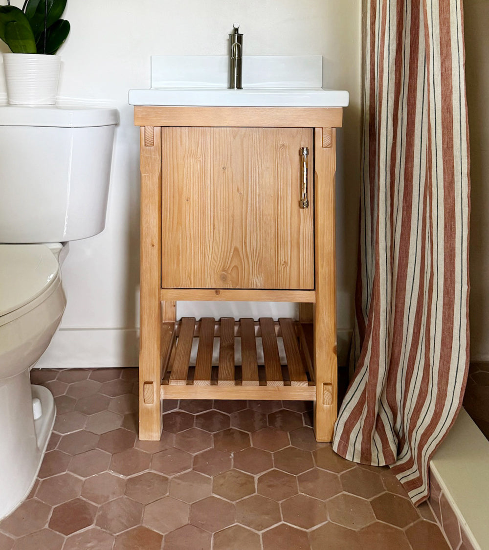 A small wooden bathroom vanity with a cabinet and slatted shelf sits beside a white toilet. The floor is covered in Desert Bloom Hex tiles by Zia Tile, and a red-and-white striped shower curtain is partially visible on the right.