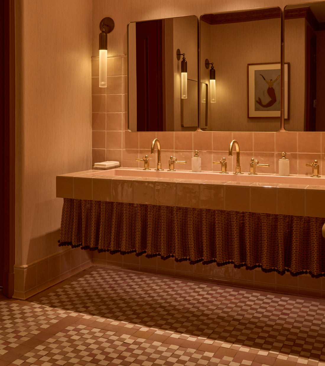 A bathroom vanity featuring a long, pink Desert Bloom zellige tile trough sink with brass faucets, a patterned fabric skirt, and Casablanca Checkerboard tile flooring.