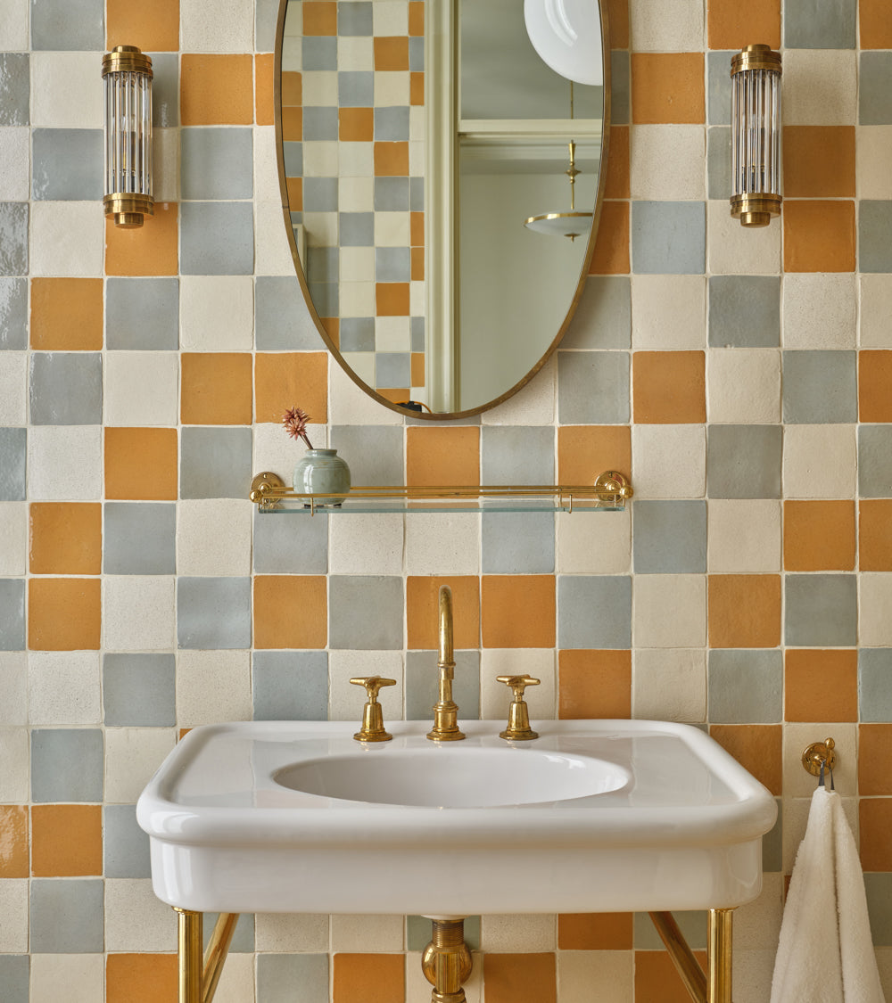 A bathroom sink with gold fixtures is set against a wall of Zia Tile’s Dorado 4x4 Square handmade tiles. Above the sink, an oval mirror and two glass sconces are mounted, with a small glass shelf holding a tiny vase.