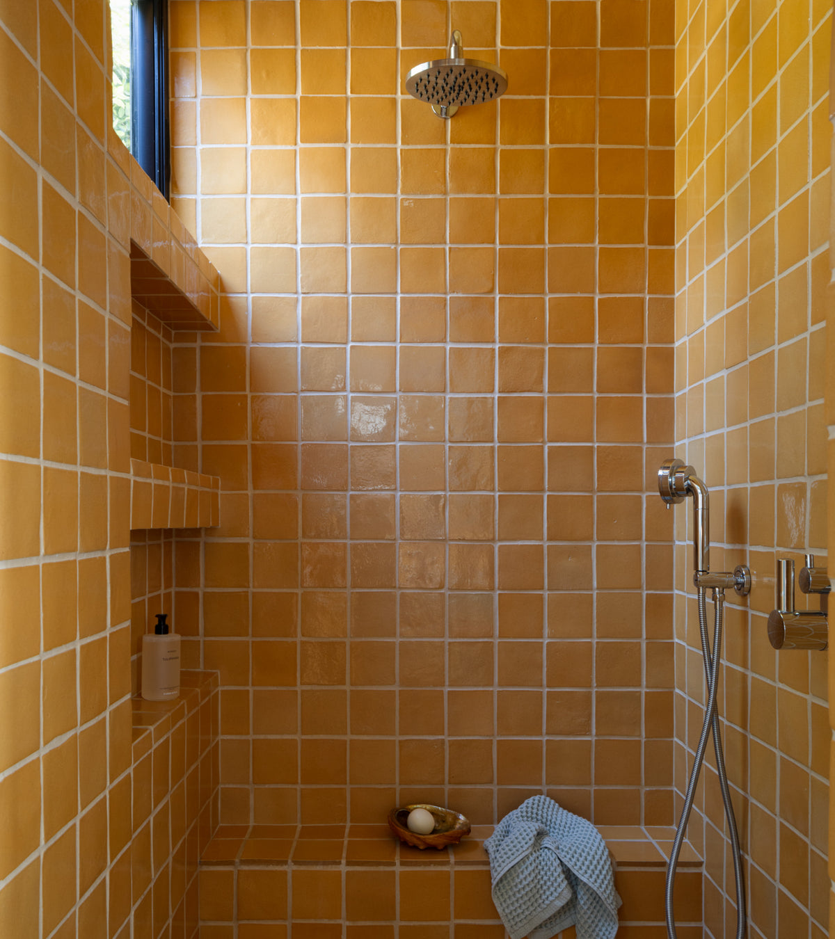 A shower featuring Dorado 4x4 Square tiles by Zia Tile in glossy orange-brown, with a rainfall and handheld showerhead, built-in shelves for soap and a bottle, a blue towel on the bench, and natural light from a small window.