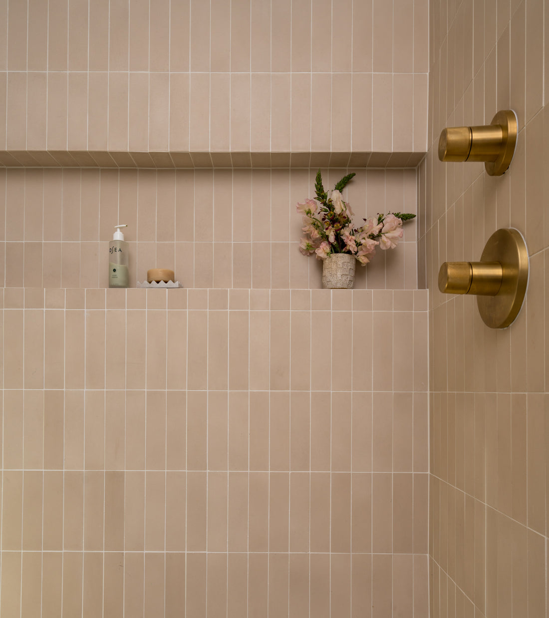 Minimalist shower featuring Zia Tile’s Dune 2x8 beige subway tile, gold shower controls, and a built-in shelf displaying a soap dispenser, bar soap, and a vase of pink flowers.