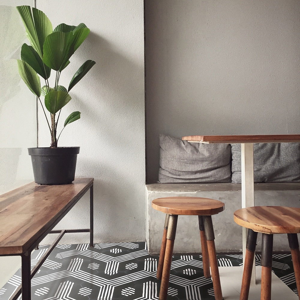 A minimalist room features a wooden bench, a potted plant, a table with two wooden stools, gray cushions on a concrete seat, and striking Echo Charcoal Hex cement flooring by vendor-unknown.