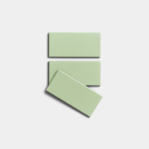 Three glossy, rectangular Eucalyptus 2x4 ceramic tiles by Zia Tile in light green are arranged on a white background—two stacked parallel and one diagonally over the bottom tile.