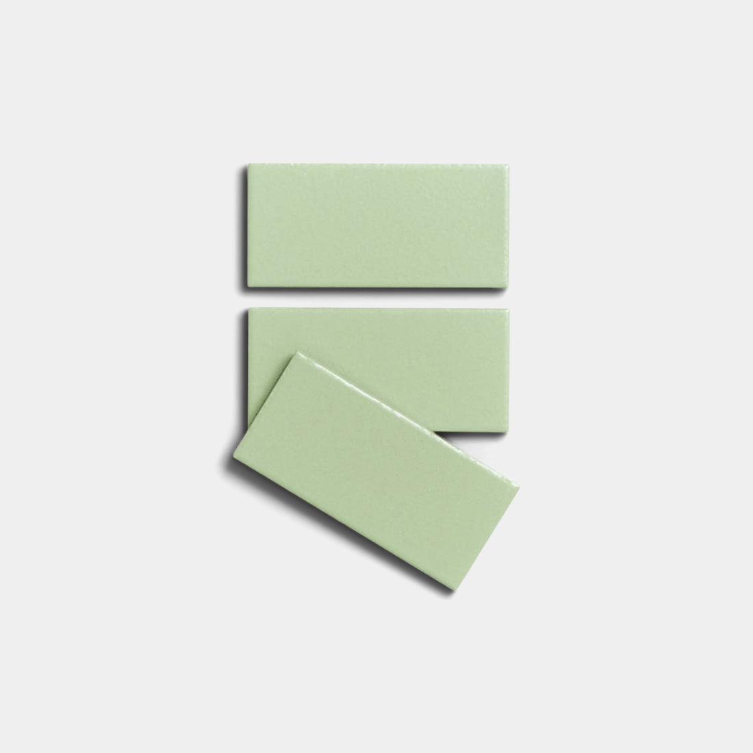 Three glossy, rectangular Eucalyptus 2x4 ceramic tiles by Zia Tile in light green are arranged on a white background—two stacked parallel and one diagonally over the bottom tile.