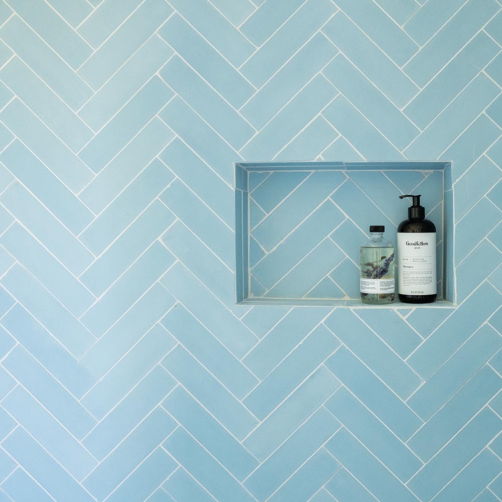 A shower wall with light blue herringbone tiles features a recessed shelf holding two bottles, one with a pump dispenser and the other with a screw cap.