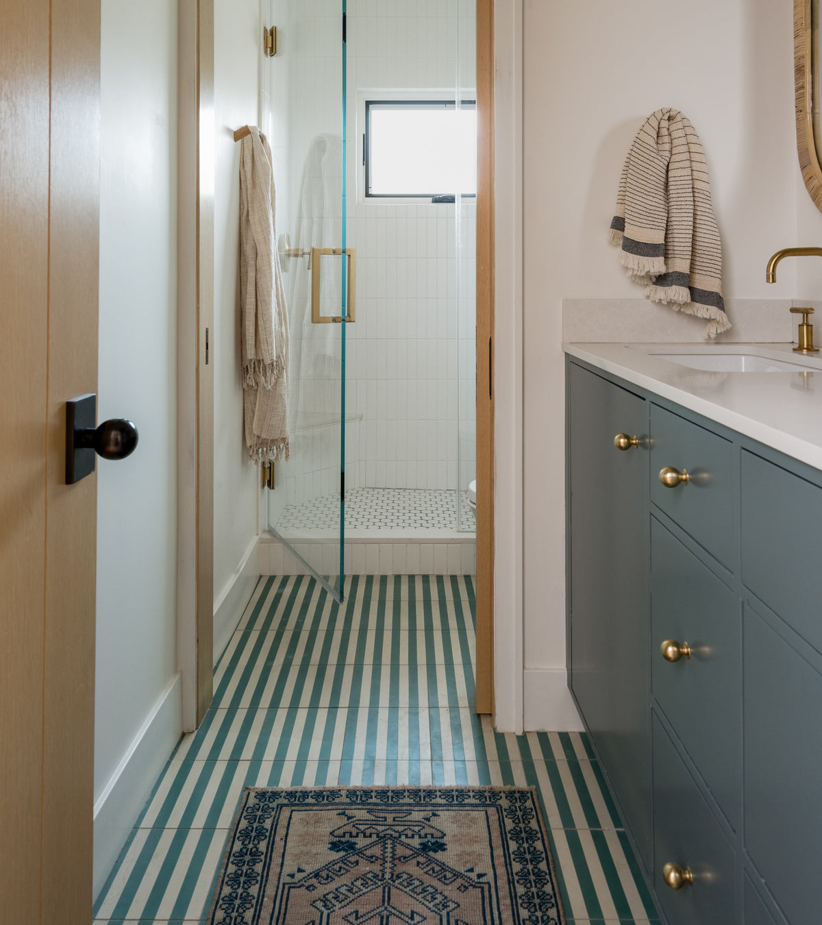 A modern bathroom featuring Zia Tile's Bateau Everglade 8x8 blue and white geometric tiles, a blue vanity with gold handles, a white countertop, glass shower enclosure, wall-mounted towel, and a patterned mat on the floor.