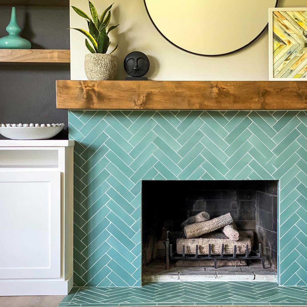 A fireplace with Everglade 2x8 ceramic tile by vendor-unknown in a herringbone pattern, a wooden mantel with decor and round mirror, plus a potted plant. White cabinetry and shelves beside the fireplace hold vases and a bowl.