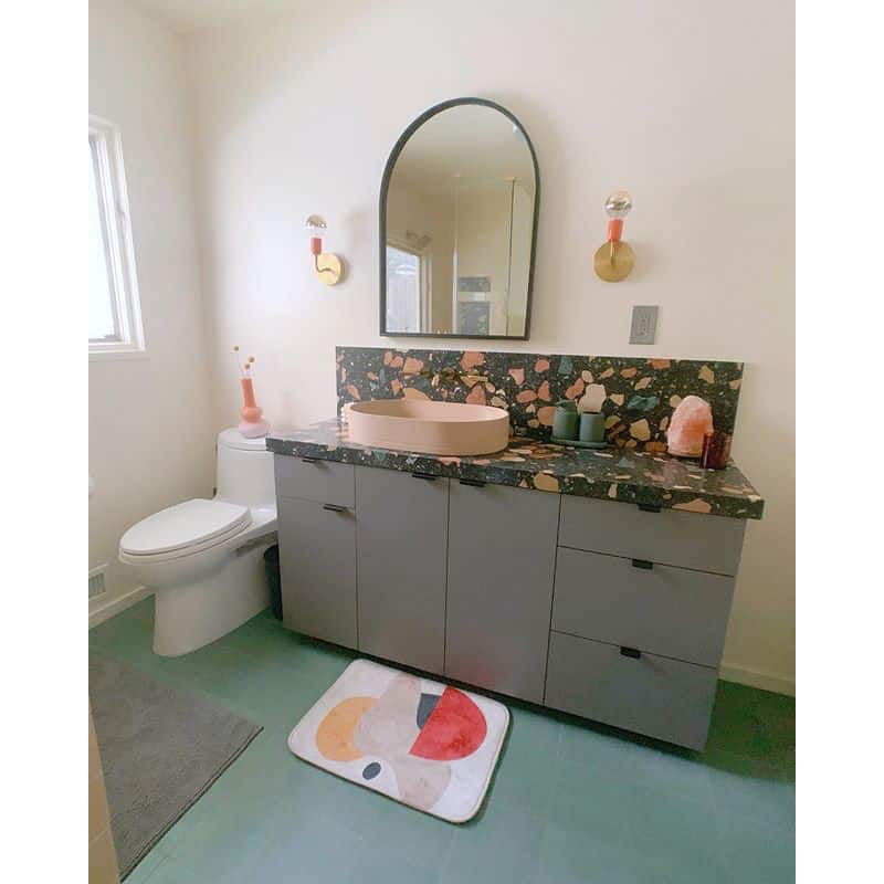 Modern bathroom featuring Everglade 2x8 tiles by vendor-unknown, terrazzo countertop, peach sink, large oval mirror, gray vanity, wall sconces, toilet, and a colorful abstract bath mat. Light blue tiles and ample natural light from a window.