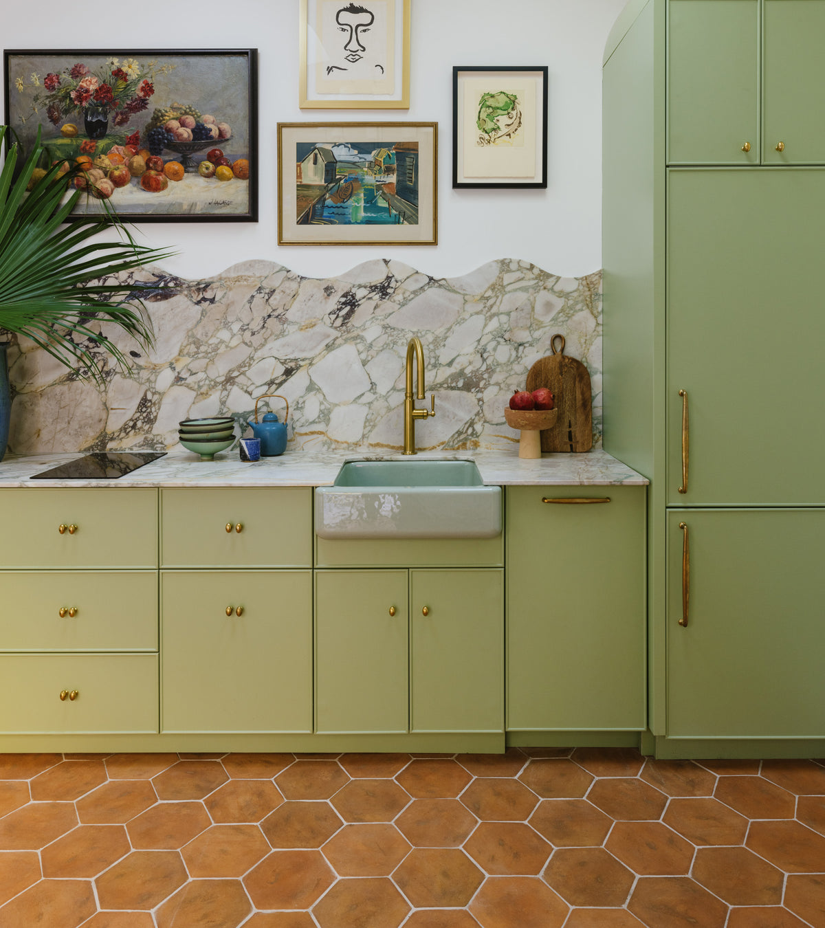 A kitchen features light green cabinets, a marble backsplash, farmhouse sink, gold faucet, and Zia Tile's 8x9 Hex + Fired Earth terra cotta floor tiles. Artworks hang above the counter with plants and kitchen items for decor.