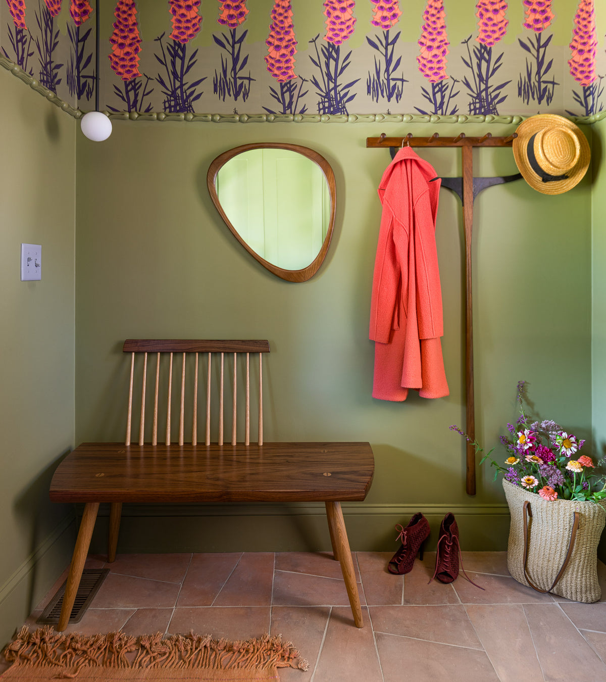 A cozy entryway with green walls, a floral border, and Toltec + Fired Earth tiles by Zia Tile features a wooden bench, mirror, coat rack with coral coat and straw hat, red shoes on the floor, and a basket of colorful flowers.