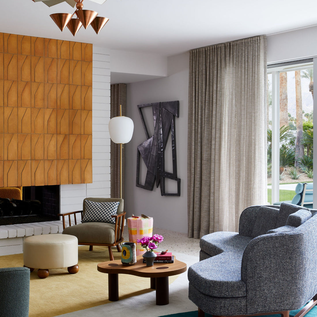 Mid-century modern living room with geometric wall panels, gray sofa, round ottoman, and a wood coffee table adorned with flowers. Abstract art hangs above neutral curtains framing a large window, while warm terra cotta accents inspired by Zia Tile’s Zocalo + Fired Earth handmade Cotto tiles from San Miguel de Allende add an inviting touch to the space.