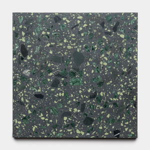 The Flagstaff 12x12 tile by Zia Tile features a dark gray background with irregular green and yellow speckles that mimic terrazzo or stone fragments, shown here on a white surface.
