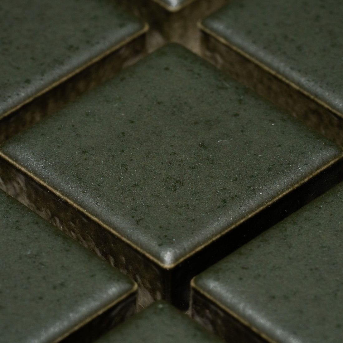 Close-up of Zia Tile's Fraser 1x1: dark green, square ceramic tiles with slightly rounded edges, visible grout lines, and a smooth matte finish featuring subtle speckles, arranged in a grid pattern.