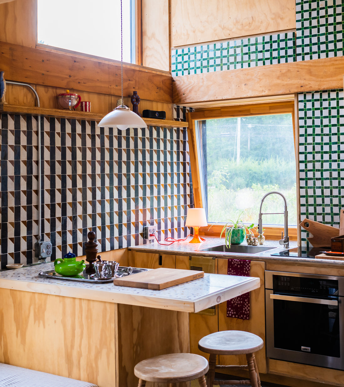 A cozy kitchen features Zia Tile's Gambit 6 Moroccan tile backsplash, wooden cabinets, a marble countertop, a stainless steel sink, and a bright window. Potted plants and kitchenware add charming decor.