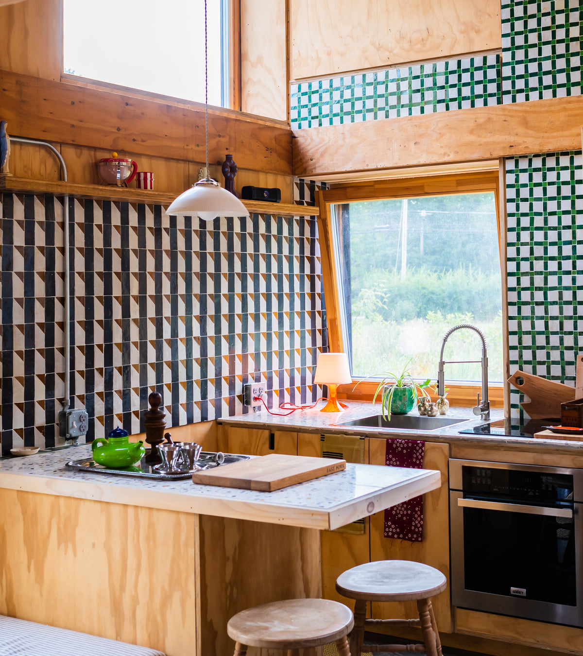 A cozy kitchen features Zia Tile's Gambit 6 Moroccan tile backsplash, wooden cabinets, a marble countertop, a stainless steel sink, and a bright window. Potted plants and kitchenware add charming decor.