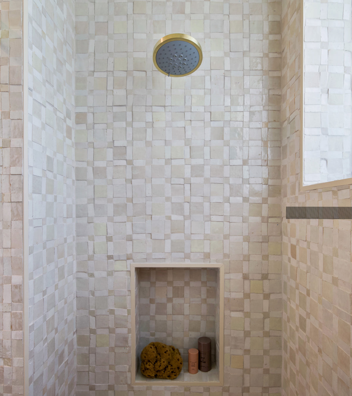 A shower featuring Zia Tile's Gambit 1 Moroccan tile in neutral tones, a gold overhead showerhead, and a built-in niche with a natural sponge and two bottles.