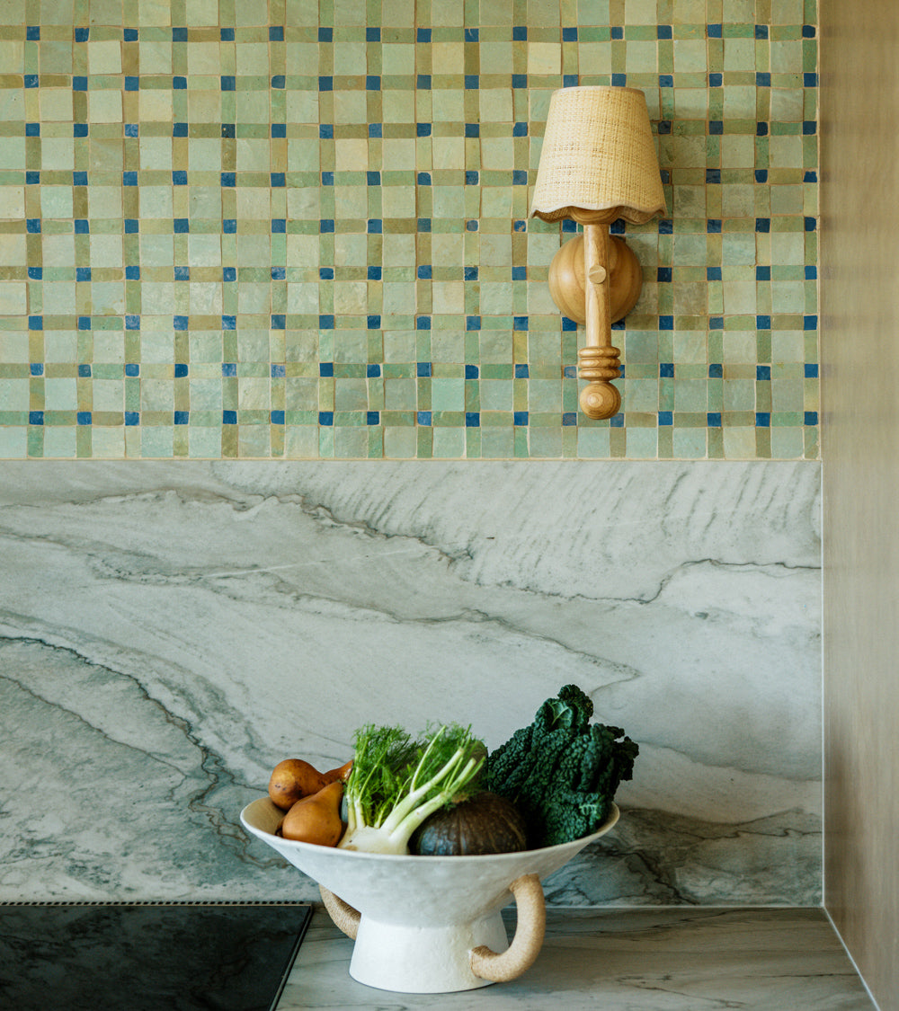 A ceramic bowl of assorted vegetables sits on a marble countertop, beneath a kitchen backsplash featuring Zia Tile's Gambit 7 mosaic zellige tiles and a wooden wall sconce with a woven lampshade.