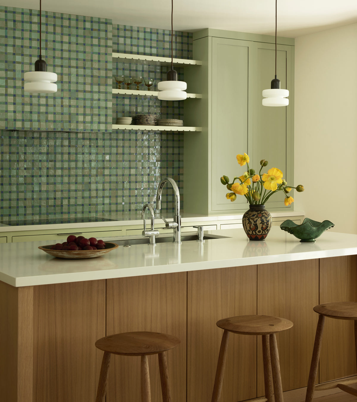 A modern kitchen features Zia Tile’s Gambit 7 green checkered Moroccan tile backsplash, a wood island with three wooden stools, light green cabinets, pendant lights, a white countertop, yellow flowers in a vase, and a bowl of apples.