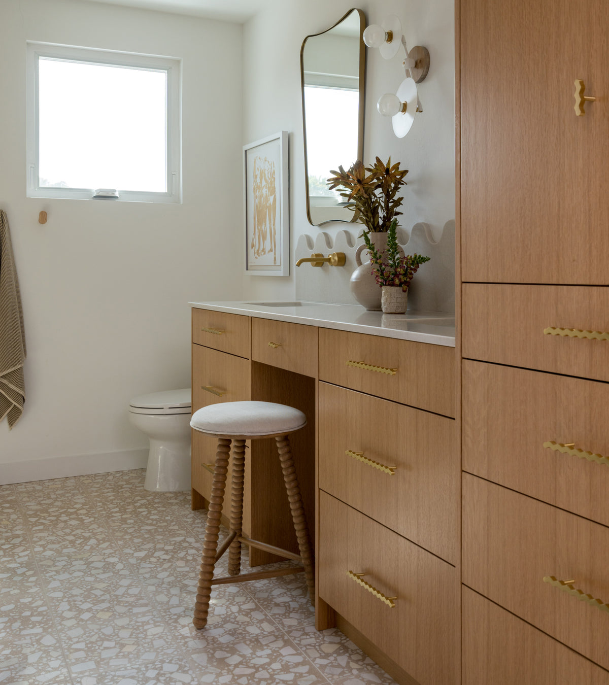 A modern bathroom with light wood cabinets, gold hardware, a white countertop, decorative mirror, wall art, flowers in a vase, a round white stool, and Zia Tile’s Gemini 12x12 Terrazzo cement tile flooring. A towel hangs near the window.