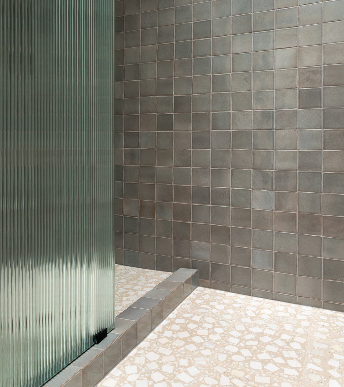 A minimalist interior showcases a wall of Zia Tile's Gemini 12x12 matte gray tiles, a step, and a floor in white and beige terrazzo cement tile. A frosted glass partition appears on the left.