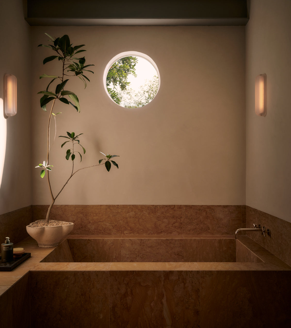 A minimalist bathroom features Giallo Reale 12x24 tan marble walls and tub by Zia Tile, a potted plant on the tub edge, handmade tile accents, a small round window with sunlight, and two soft wall lights flanking the space.