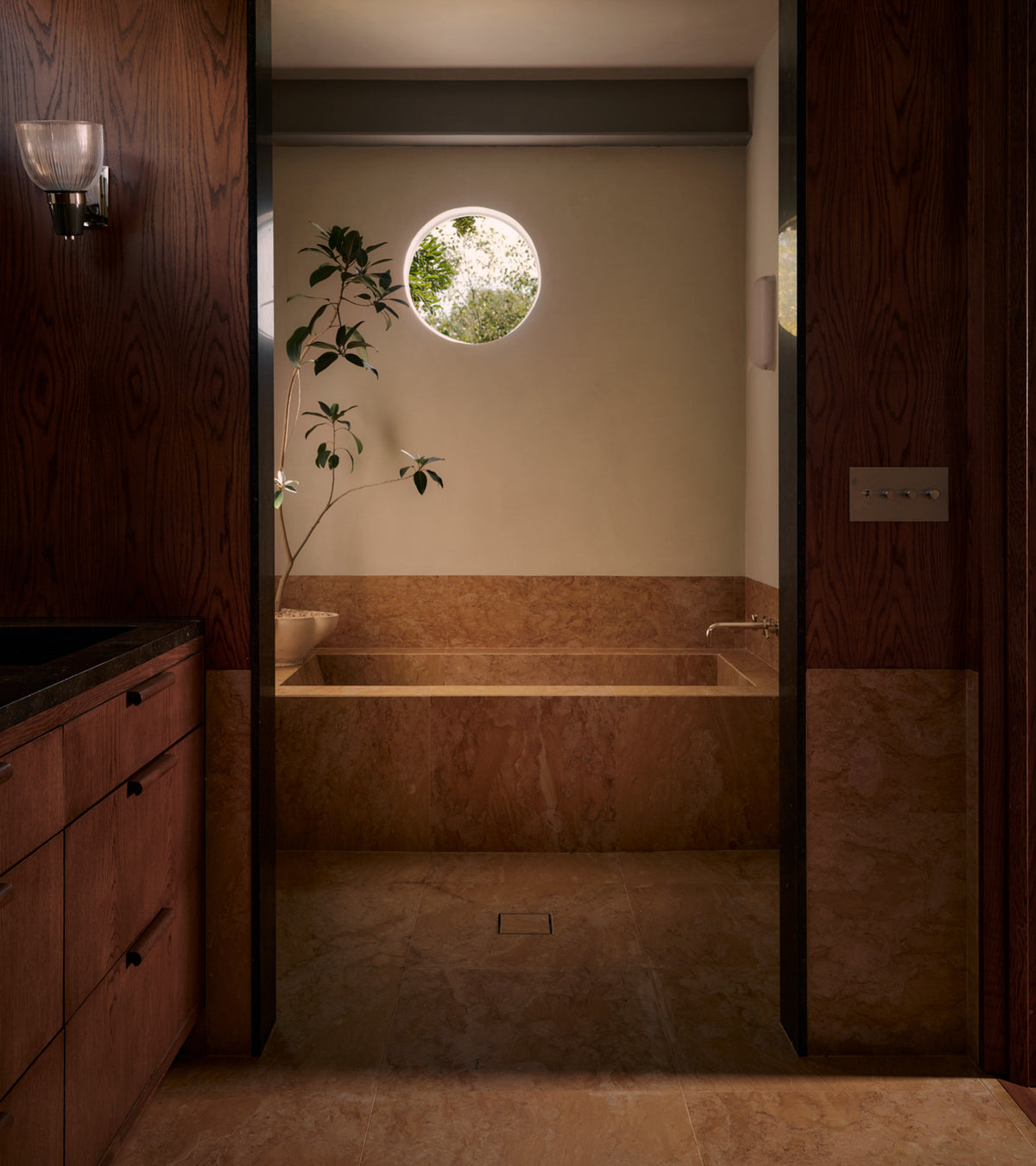 A minimalist bathroom features a beige marble bathtub and floor, Giallo Reale 24x24 tiles from Zia Tile as handmade accents, a small plant, wood-paneled walls, and a round window bringing in natural light.
