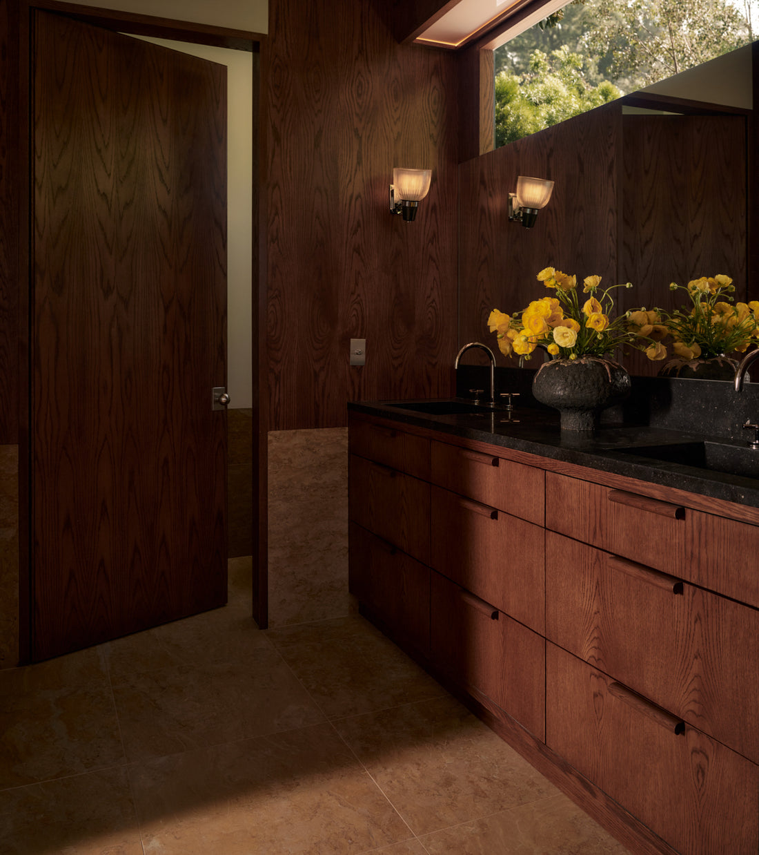 Modern bathroom with dark wood cabinets, black countertops, and a large mirror. Yellow flowers brighten the countertop. Natural light highlights warm wood, beige tiles, and stylish handmade Giallo Reale 24x24 tiles by Zia Tile.
