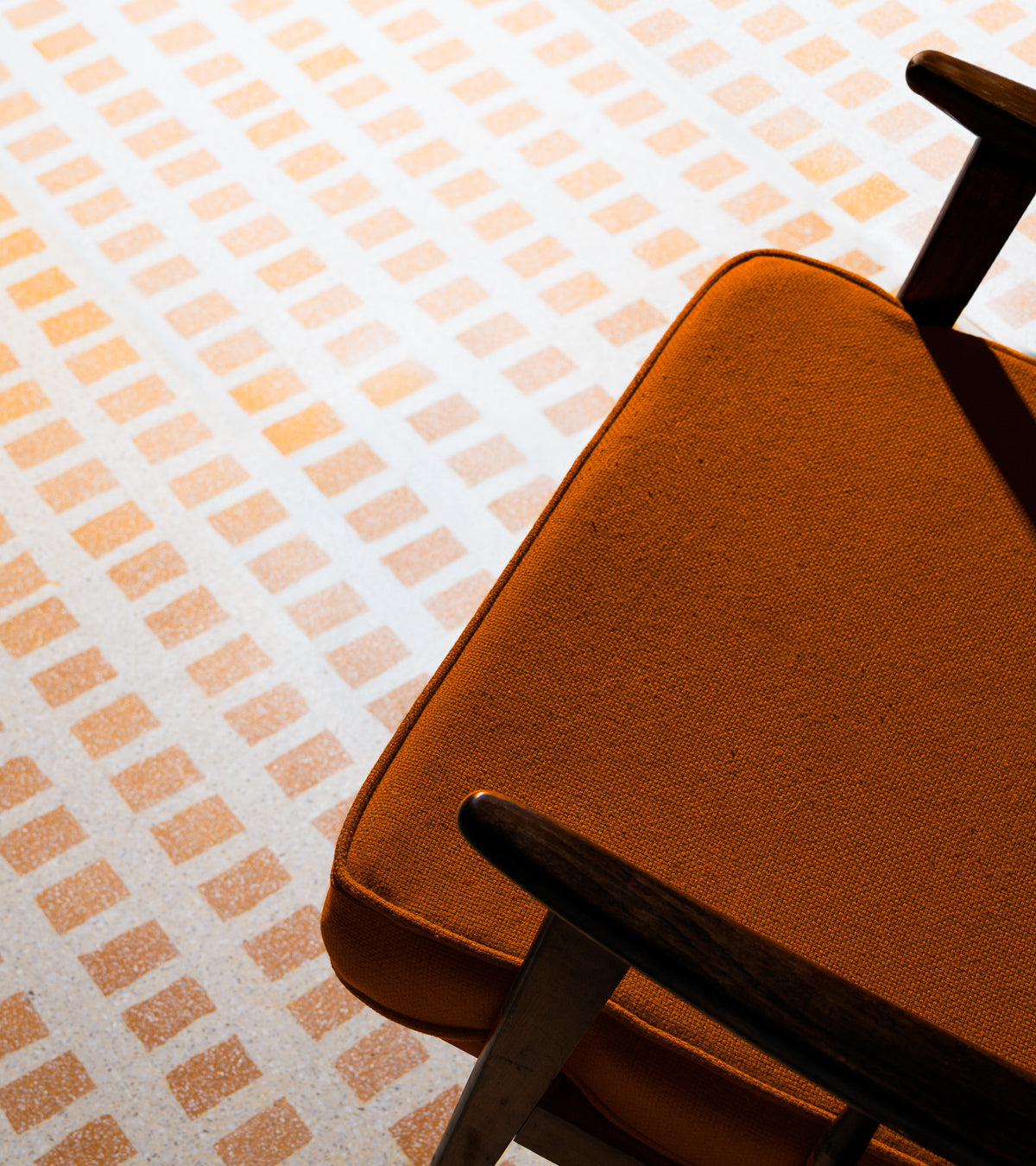 A close-up of an orange upholstered chair with wooden armrests sits on a floor featuring Zia Tile’s Constellation Bone + Ginger 24x24 tiles, showcasing a retro geometric pattern of orange and white squares reminiscent of classic Terrazzo.