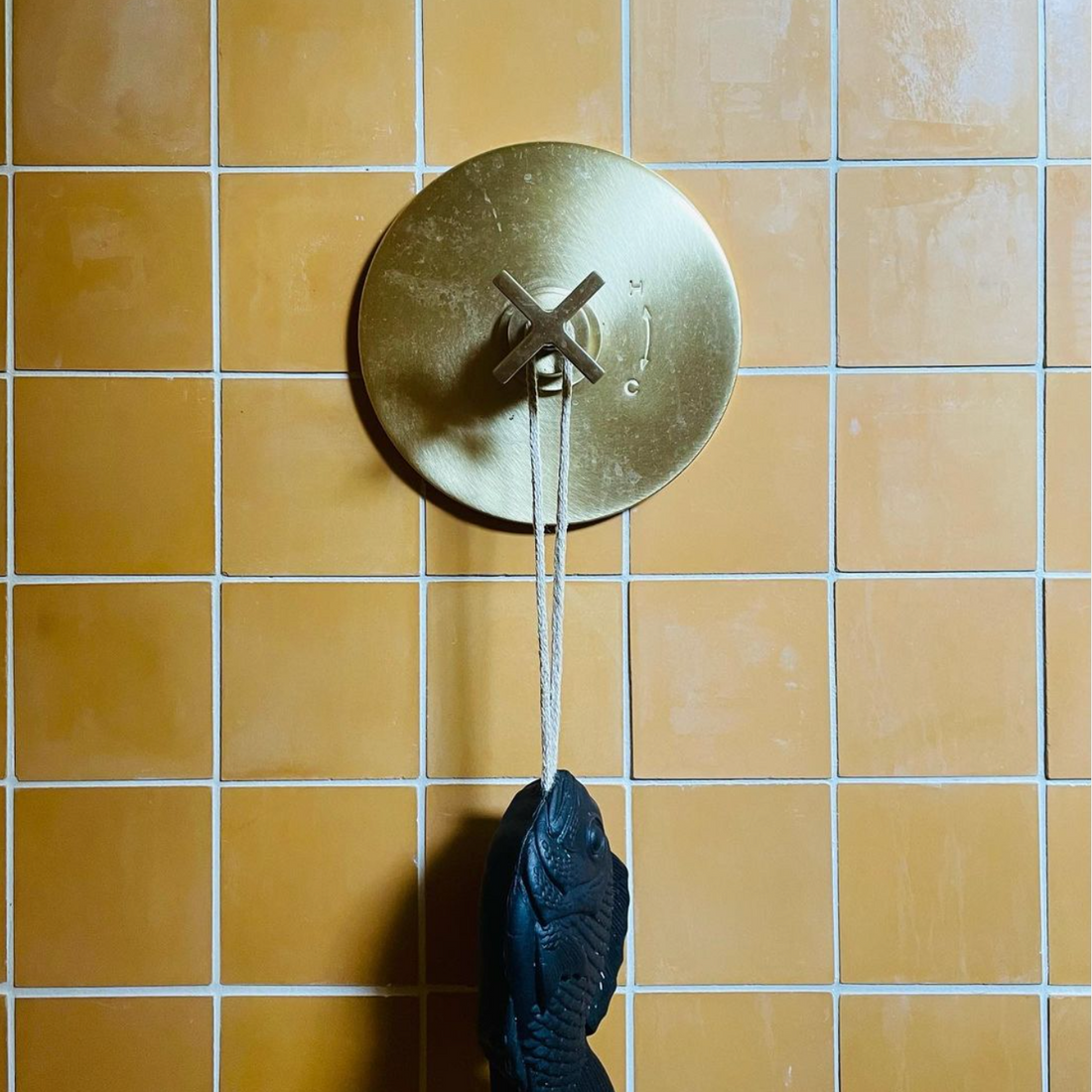 A gold shower control with a round handle is mounted on a vendor-unknown Ginger 4x4 orange tile wall, and a black fish-shaped object hangs from a rope on the handle.