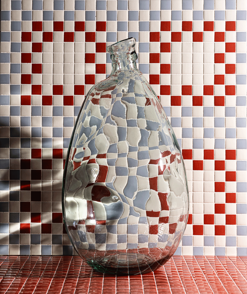 A clear, distorted glass bottle stands in front of a wall with red, blue, and white mosaic tiles arranged in a pattern that includes two red heart shapes. The glass creates a wavy visual effect on the tiles behind it.