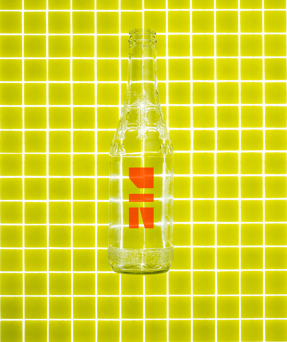 A clear glass bottle with a bold red geometric design stands upright on a bright yellow grid-patterned background, creating a modern and vibrant visual contrast.