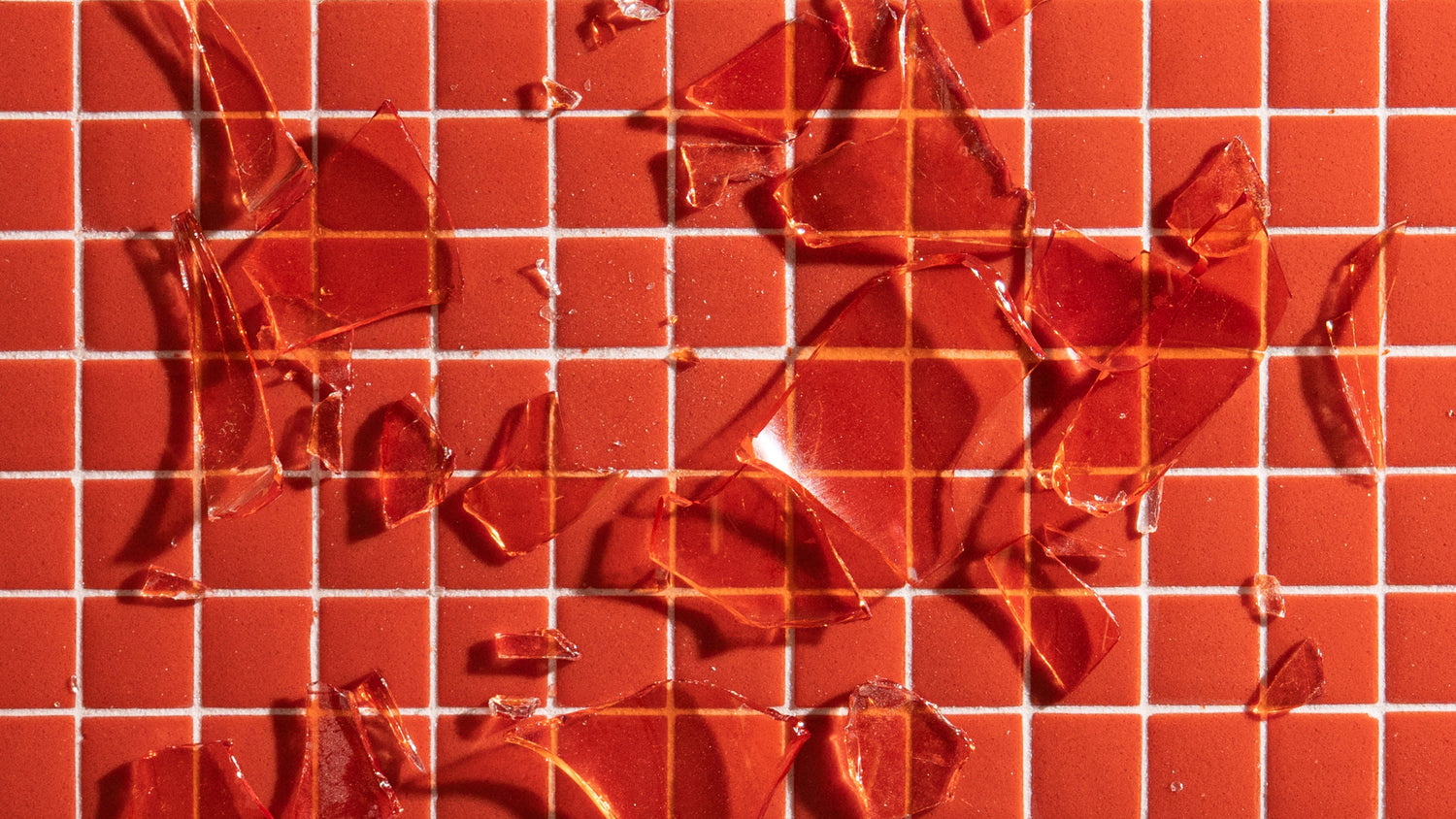 Broken pieces of reddish glass scattered across a grid of small, square, red tiles with white grout lines. The sharp shards contrast with the clean, geometric tile pattern underneath.