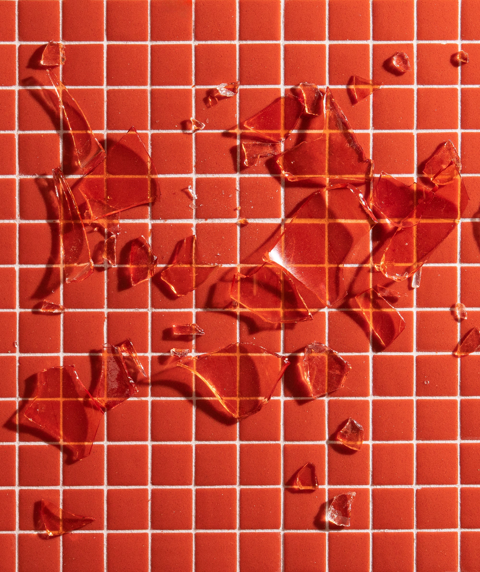 Shards of broken red glass scattered across a red tiled surface with white grout lines, forming an irregular pattern.