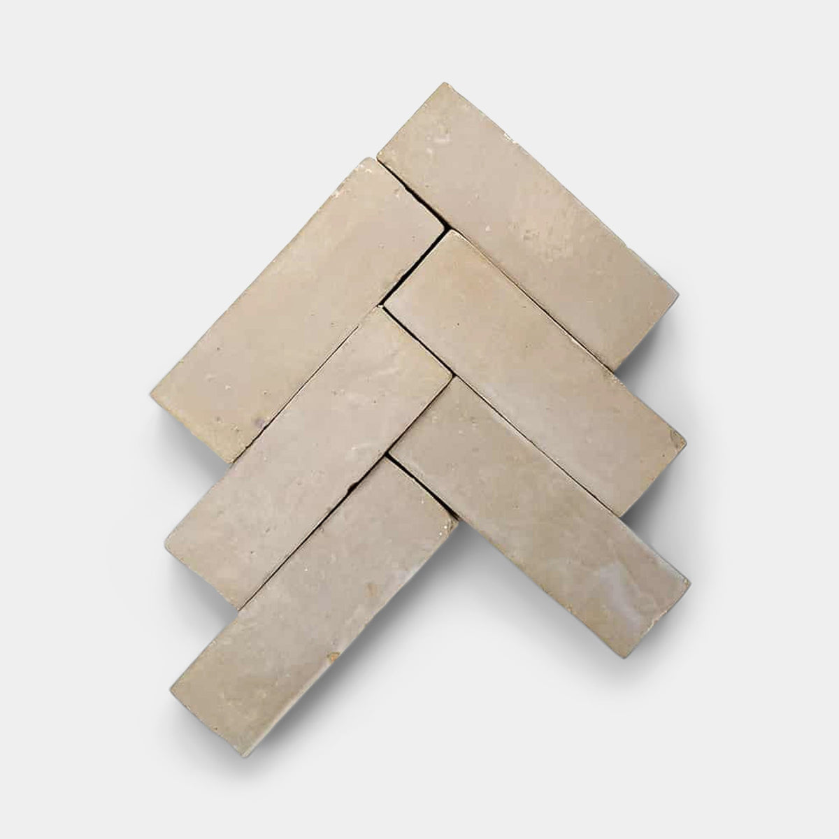 Six beige Glazed Earth 2x2 tiles by Zia Tile are arranged in a herringbone pattern on a light gray background, showcasing visible grout lines and subtle surface texture for a geometric, symmetrical look.